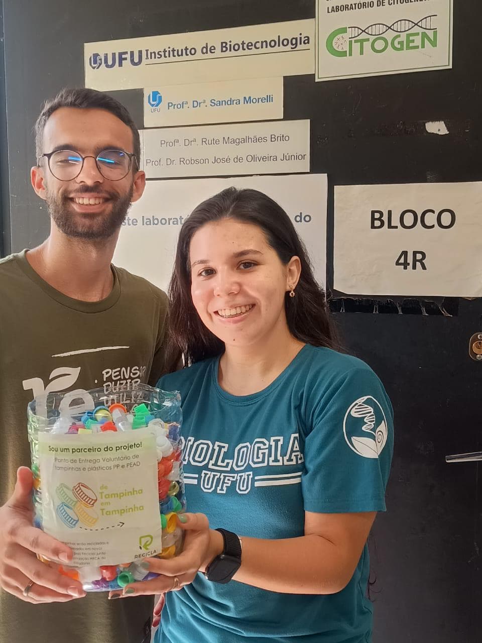 Parceiros do ponto de coleta no Instituto de Biotecnologia da UFU segurando um recipiente cheio de tampinhas