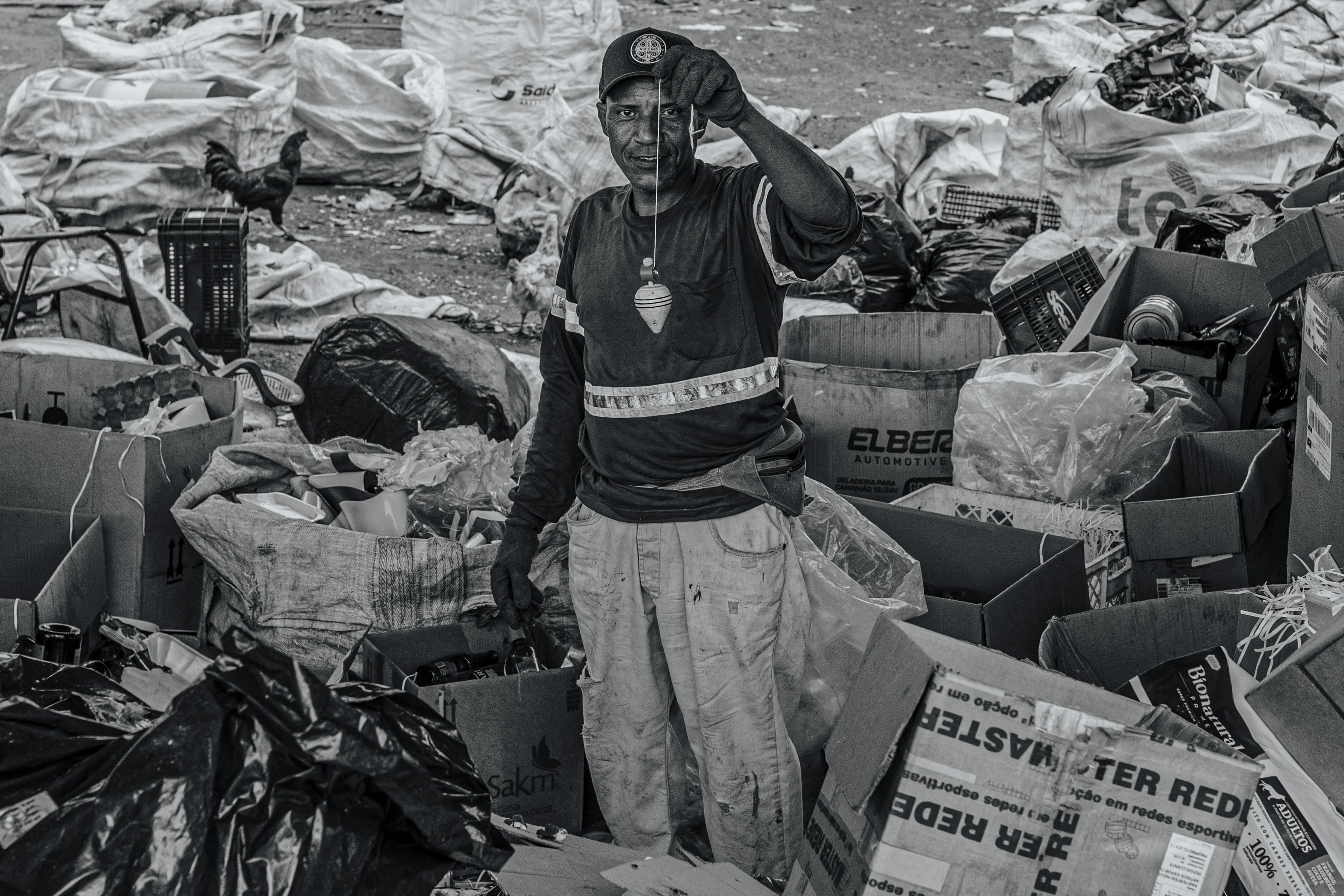 Catador em um pátio de triagem de recicláveis ergue um pião de plástico reciclado, olhando para a câmera